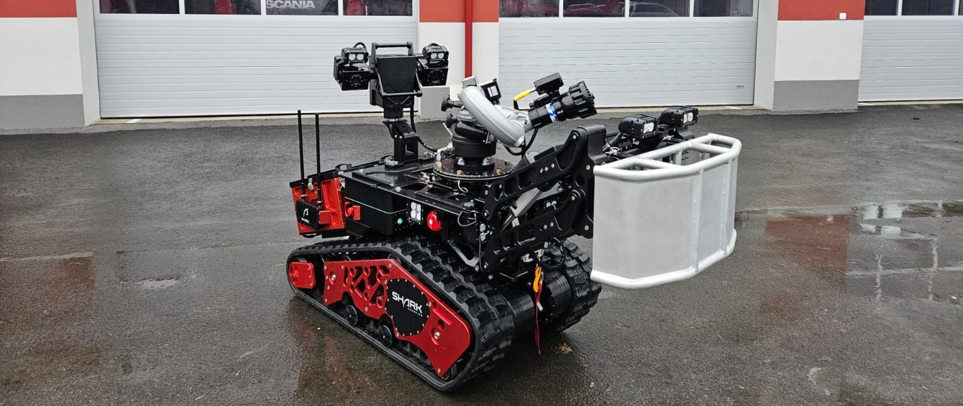 Zdjęcie przedstawia zdalnie sterowany pojazd do gaszenia pożarów i likwidacji zagrożeń CBRNE znajdujący się przed bramą garażową Jednostki Ratowniczo Gaśniczej w Nowej Sarzynie.