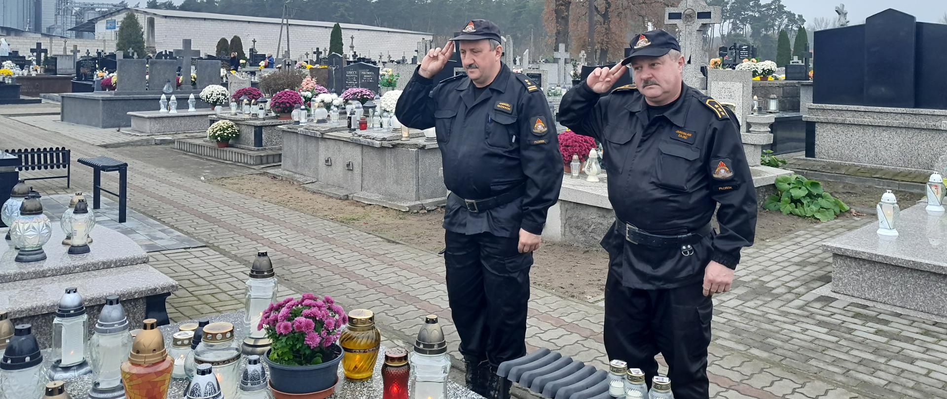 Dwóch strażaków w czarnych ubraniach dowódczo-sztabowych oddaje honor na cmentarzu, przed grobem na którym ustawione są znicza oraz kwiaty.