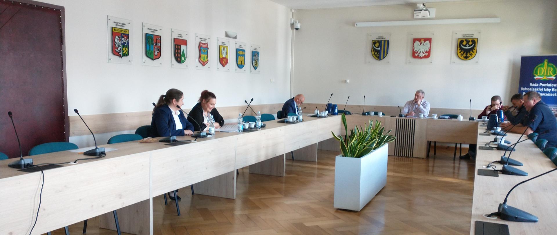 W jasnej sali konferencyjnej odbywa się spotkanie kilku osób siedzących przy stołach ustawionych w kształt podkowy. Na stołach znajdują się mikrofony i dokumenty, a na ścianach wiszą herby miast i instytucji. W centrum pomieszczenia stoi donica z rośliną
