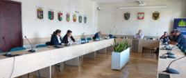 W jasnej sali konferencyjnej odbywa się spotkanie kilku osób siedzących przy stołach ustawionych w kształt podkowy. Na stołach znajdują się mikrofony i dokumenty, a na ścianach wiszą herby miast i instytucji. W centrum pomieszczenia stoi donica z rośliną