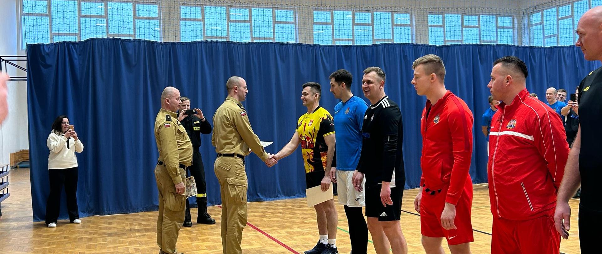 W hali sportowej dwóch mężczyzn w mundurach uściska sobie dłonie, podczas gdy inni mężczyźni w strojach sportowych stoją w rzędzie. Wydarzenie jest częścią ceremonii wręczenia nagród.