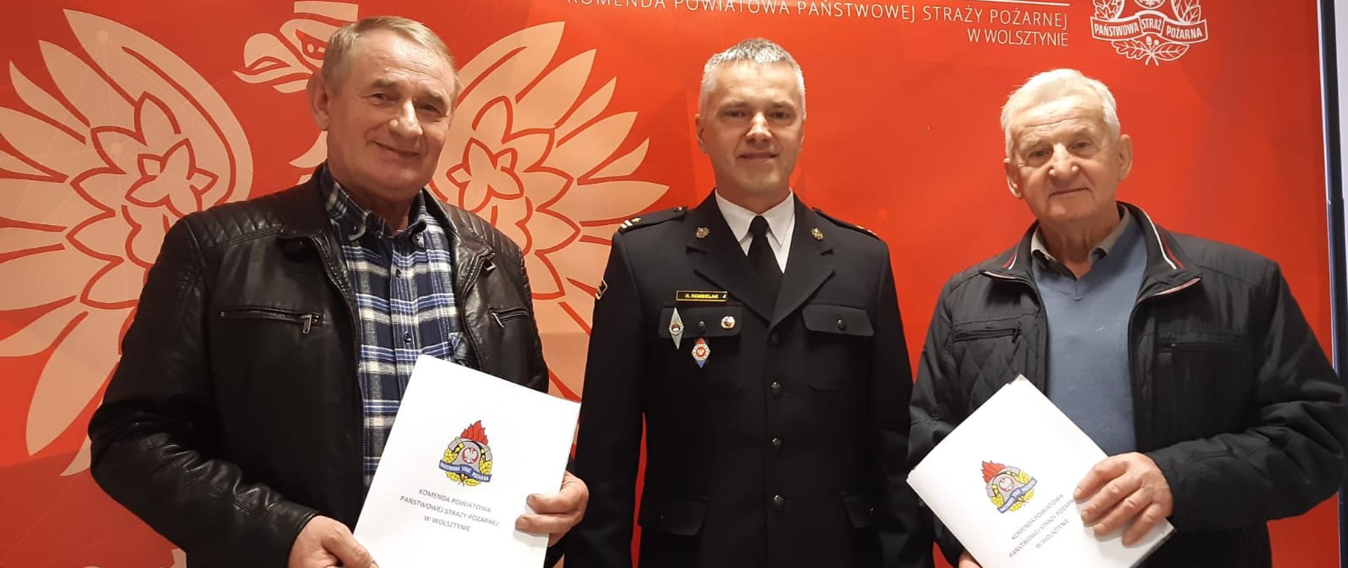 Komendant Powiatowy PSP stoi z druhami OSP. Druhowie trzymają w dłoniach teczki z decyzjami o przyznaniu świadczenia.