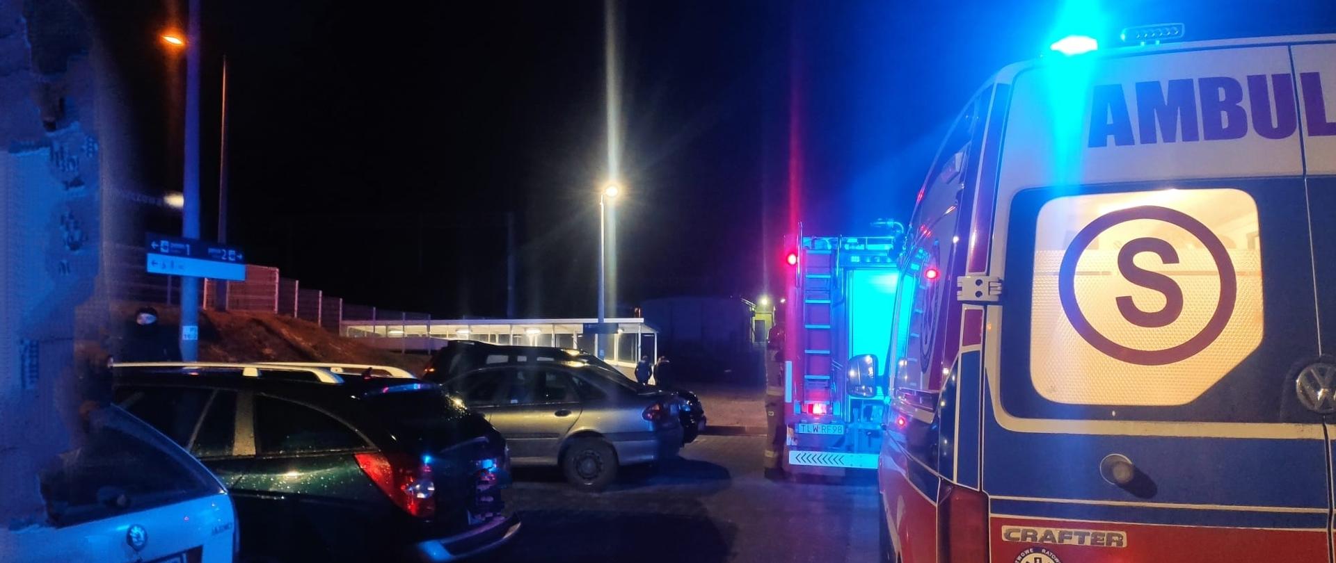Pora nocna, na pierwszym panie tył karetki pogotowia oraz samochody zaparkowane na parkingu obok dworca, nieco dalej samochód ratowniczo-gaśniczy, w oddali pociąg stojący na peronie 