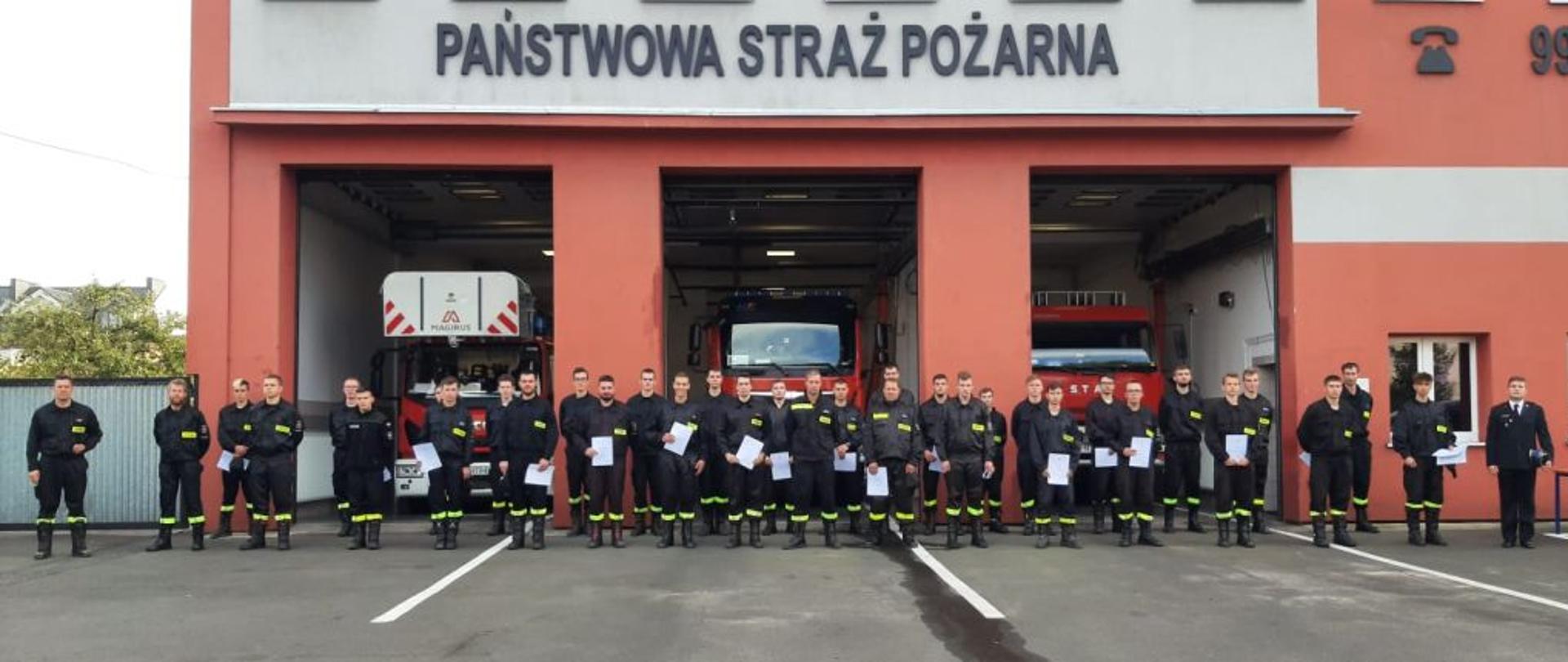 Dzień. Na tle budynku Komendy Powiatowej oraz samochodów strażackich w otwartych bramach garażowych stoi grupa strażaków w obecności Zastępcy Komendanta Powiatowego PSP. W rekach trzymają zaświadczenia o ukończeniu szkolenia.