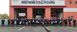 Dzień. Na tle budynku Komendy Powiatowej oraz samochodów strażackich w otwartych bramach garażowych stoi grupa strażaków w obecności Zastępcy Komendanta Powiatowego PSP. W rekach trzymają zaświadczenia o ukończeniu szkolenia.