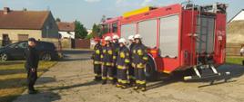 Plac przed siedzibą OSP Jutrosin. Na nim wóz strażacki. Z jego lewej strony sześciu strażaków w ubraniach specjalnych. Na głowach maja hełmy. Przed strażakami zastępca komendanta powiatowego PSP w Rawiczu