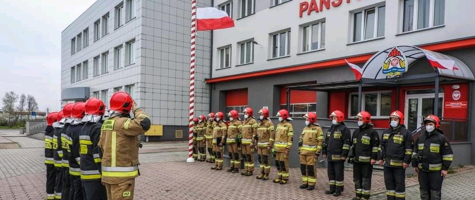 na zdjęciu widać strażaków w czasie uroczystej zmiany służby, podczas której podnoszona jest flaga RP na masz, strażacy stoją w dwóch grupach na przeciwko siebie, na środku dowódca zmiany stojący tyłem, po tle budynek jednostki ratowniczo gaśniczej 