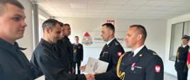 Strażak w mundurze składa gratulacje drugiemu strażakowi. Wręczane są dokumenty.