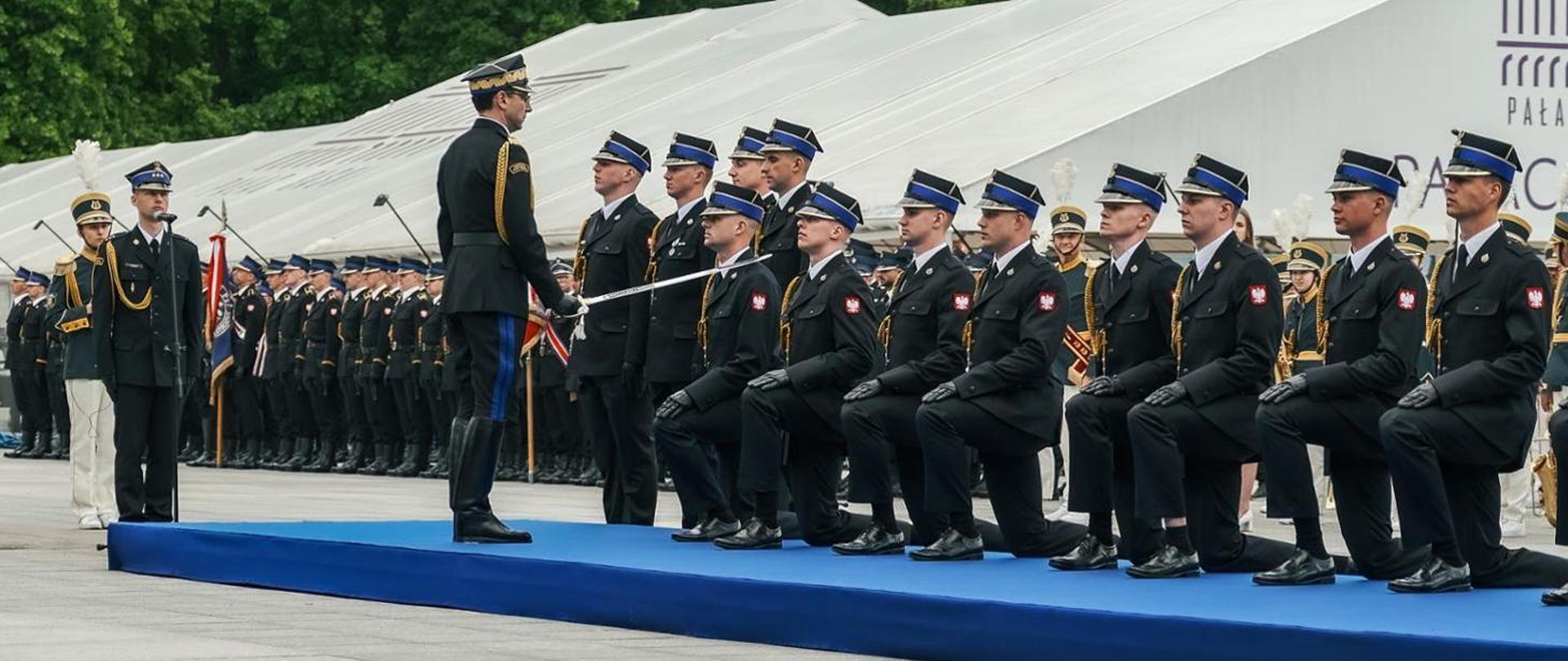 Na zdjęciu przedstawiono uroczystą ceremonię pasowania na pierwszy stopień oficerski funkcjonariuszy Państwowa Straż Pożarna. Uroczystość odbywa się podczas oficjalnych obchodów z udziałem licznie zgromadzonych pododdziałów strażackich ustawionych w szyku za główną sceną.
Na podwyższeniu przykrytym niebieskim podestem klęczy kilku funkcjonariuszy w galowych mundurach PSP z charakterystycznymi rogatywkami z niebieskim otokiem. Przed nimi stoi przełożony w mundurze galowym, który dokonuje aktu symbolicznego pasowania szablą. Gest ten oznacza nadanie stopnia oficerskiego oraz przyjęcie do korpusu oficerów Państwowej Straży Pożarnej.
W tle widoczne są kolejne pododdziały strażaków ustawione w zwartym szyku, poczty sztandarowe oraz orkiestra. Całość odbywa się w podniosłej atmosferze, podkreślającej rangę wydarzenia oraz znaczenie tradycji i ceremoniału służb mundurowych.