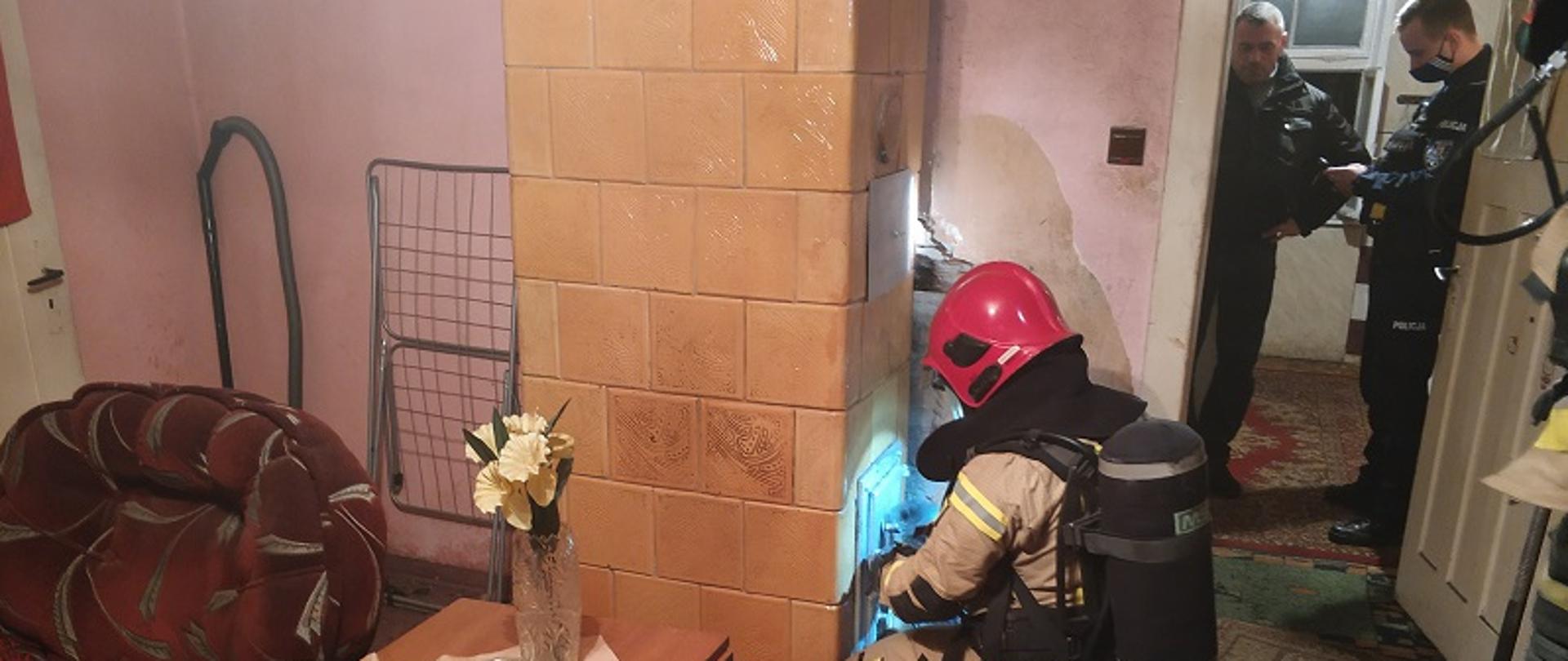 Zdjęcie wykonane w pokoju mieszkalnym. W centralnej części zdjęcia żółty piec kaflowy, a przed nim z prawej strony klęczący strażak w piaskowym ubraniu specjalnym i z butlą aparatu powietrznego na plecach. W otworze drzwiowym po prawej stronie stoją dwaj policjanci w służbowych mundurach