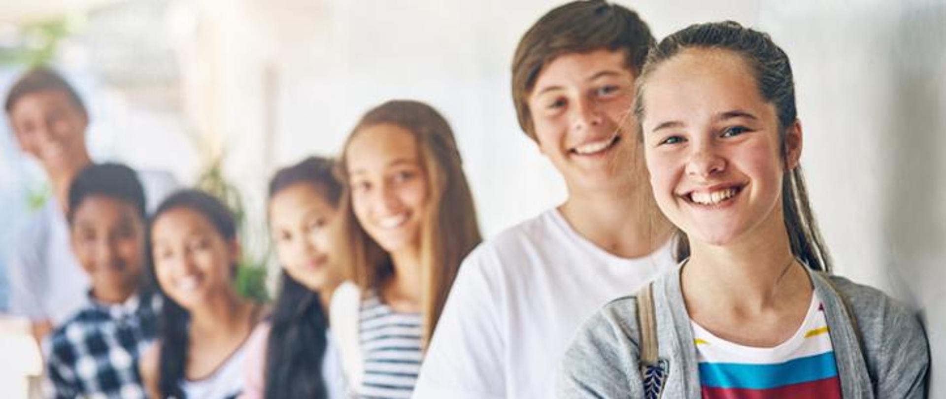 Grupa uśmiechniętej młodzieży stojąca w rzędzie w jasnym korytarzu. Na pierwszym planie nastoletnia dziewczyna z plecakiem, patrząca z radością w stronę obiektywu. 