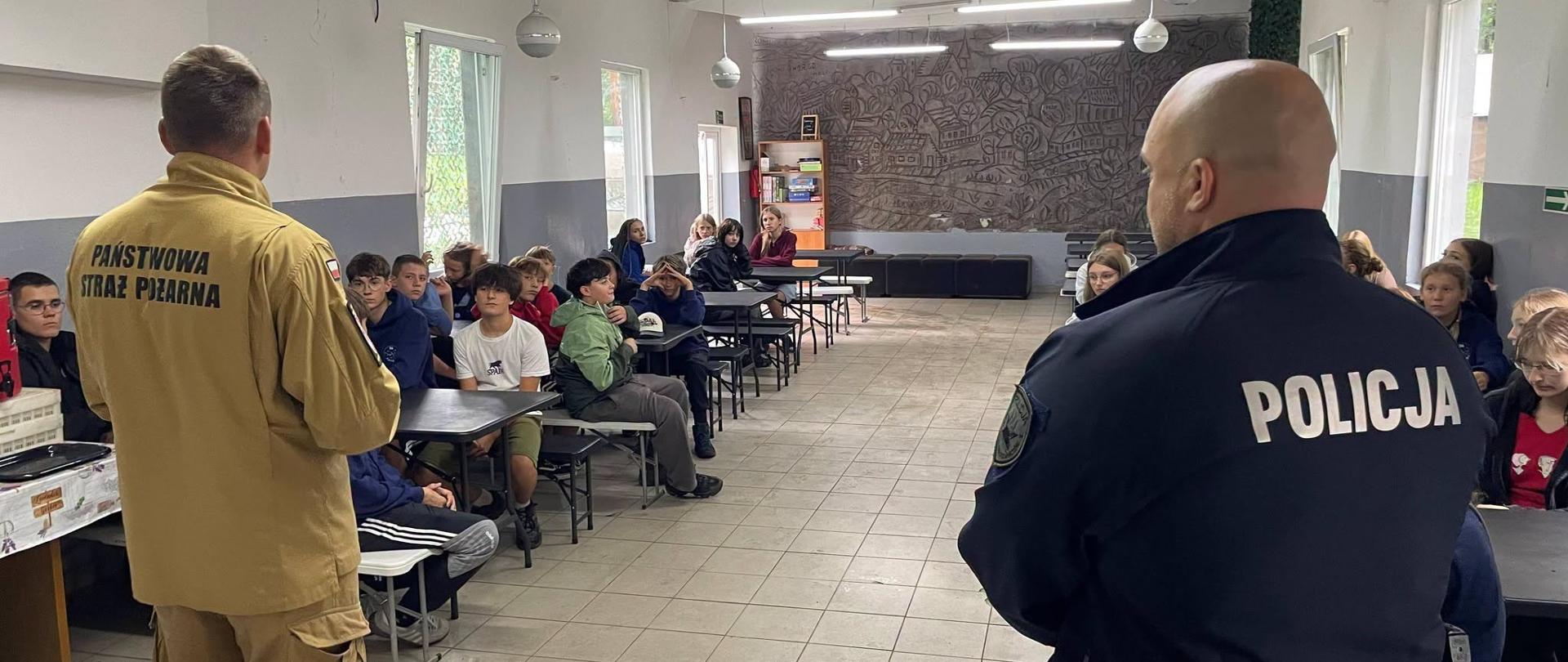 Na zdjęciu widać spotkanie profilaktyczno-edukacyjne z udziałem strażaka Państwowej Straży Pożarnej i policjanta. Funkcjonariusze stoją przed grupą dzieci i młodzieży siedzących przy stołach w dużej sali. Strażak ubrany jest w piaskowy mundur z napisem „Państwowa Straż Pożarna”, a policjant w granatowy mundur z dużym napisem „Policja” na plecach. Uczestnicy spotkania uważnie słuchają prowadzących.