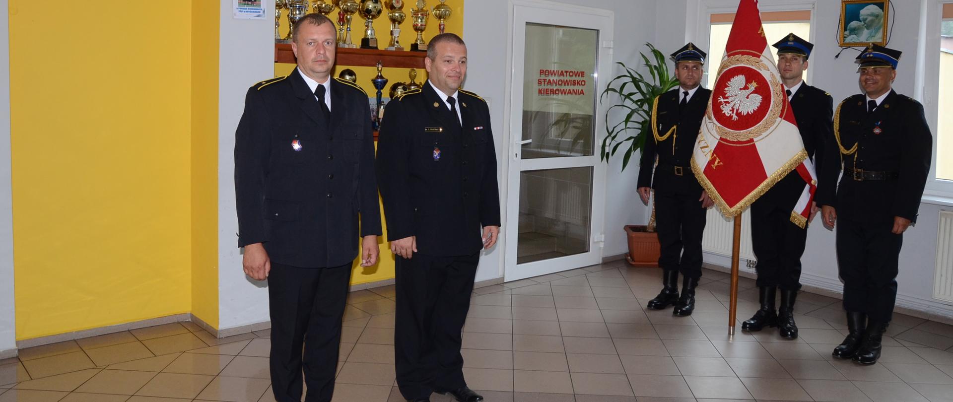 Sala w komendzie straży. Dwóch strażaków wyznaczonych do awansu. W tle poczet sztandarowy.