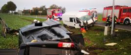 Dwa uszkodzone samochody dostawcze , czarny pojazd leży na boku, na polu, mocno uszkodzony, biały pojazd stoi na poboczu z uszkodzonym przodem, w tle widać dwa pojazdy straży pożarnej i karetkę pogotowia oraz kilku strażaków