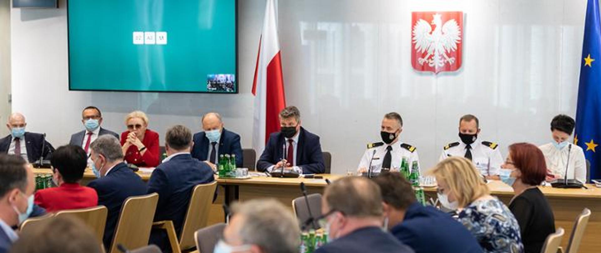 Na zdjęciu przy stołach siedzą uczestnicy posiedzenia tj. członkowie parlamentarnego zespołu strażaków oraz wiceminister SWiA, komendant główny PSP wraz z zastępcą, na ścianie na wprost wisi godło Polski, po lewej stronie ekran z niebieskim tłem