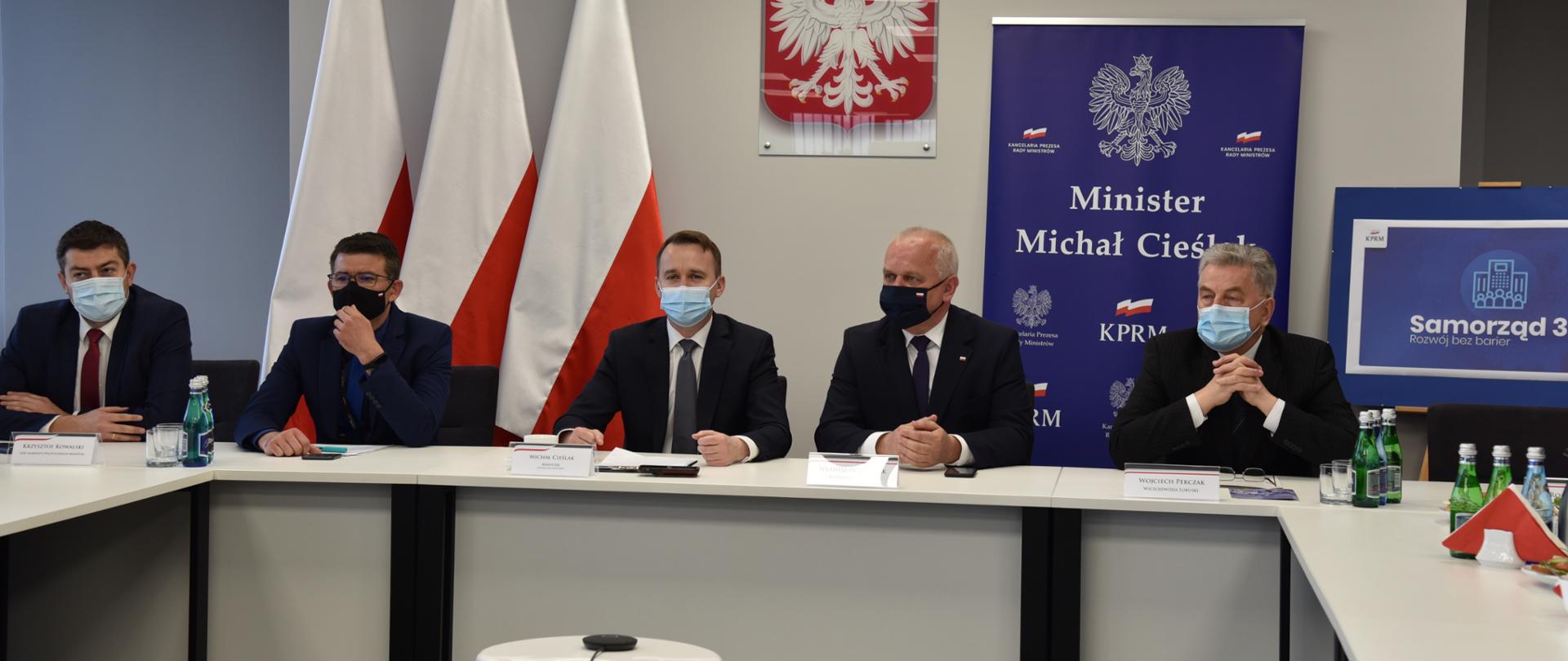 Przy białym stole prezydialnym siedzą: doradca ministra, dyrektor Biura Wojewody, minister Cieślak, Wojewoda Dajczak, Wicewojewoda Perczak. Za nimi biało-czerwone flagi, gosło i ścianka promocyjna ministra 