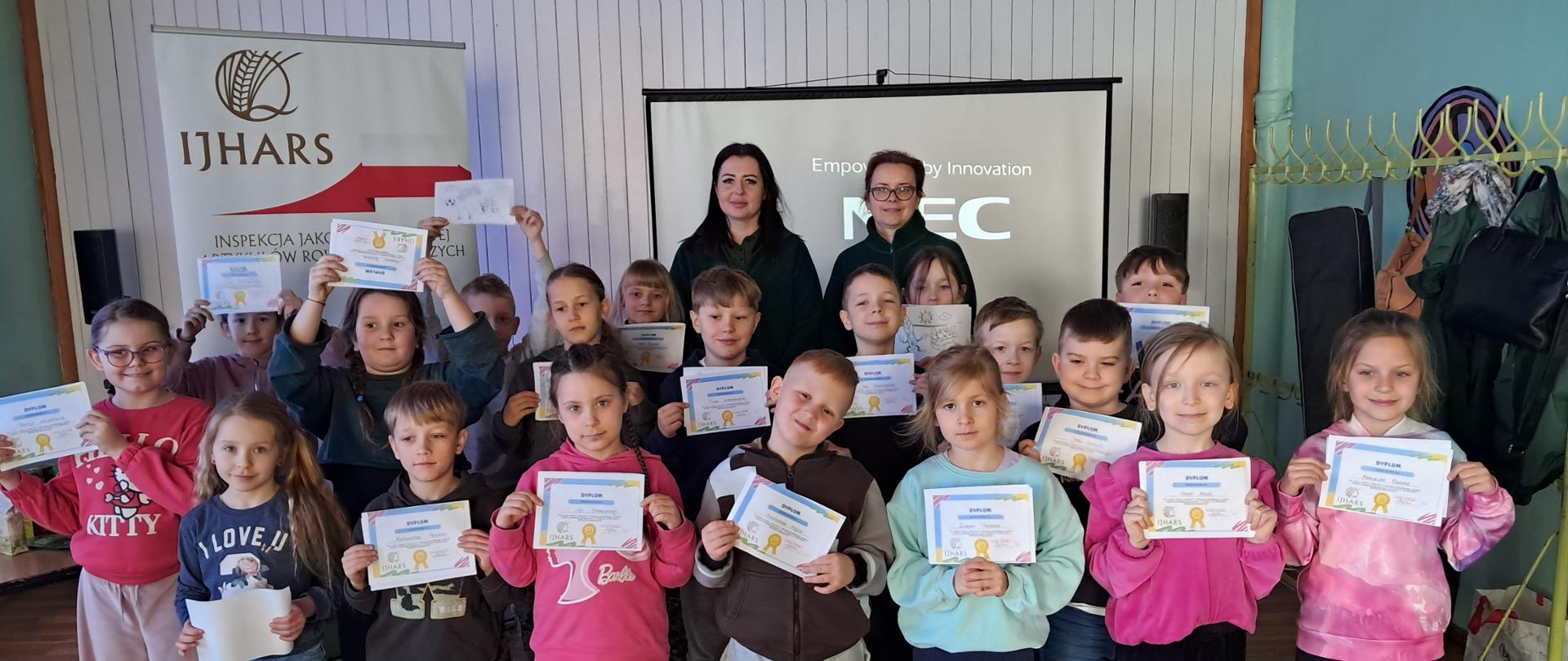Zdjęcie grupowe po zajęciach edukacyjnych: uśmiechnięte dzieci stoją razem i trzymają w rękach dyplomy. W tle widoczne jest logo IJHARS, a obok grupy stoją dwie inspektorki Inspekcji, które towarzyszyły zajęciom.