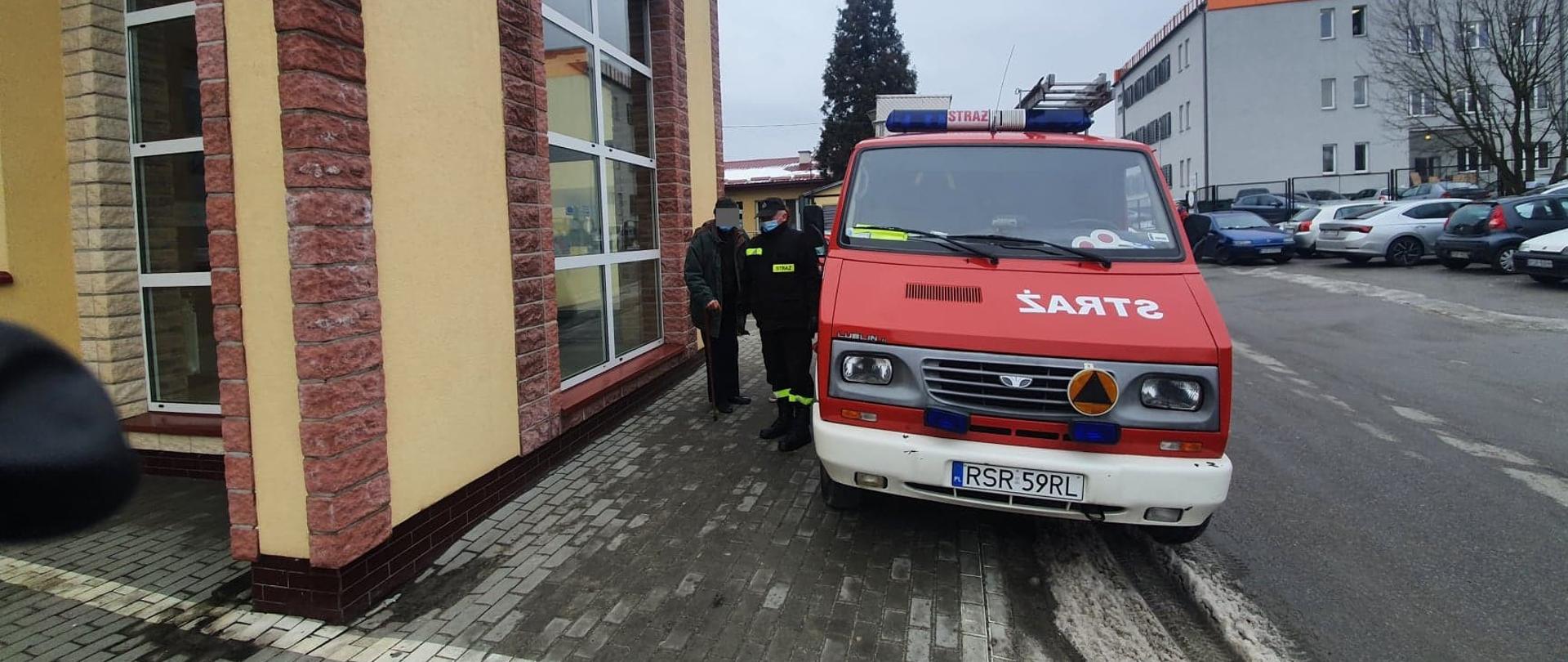 Na zdjęciu w centralnej jego części samochód pożarniczy. Po lewej stronie strażak w umundurowaniu koszarowym asystujący osobę przewożoną do szczepienia. Po lewej stronie budynek z żółtą elewacją z wykończeniem klinkierowym. W tle po prawej stronie szary budynek Poczty Polskiej oraz widoczne samochody na parkingu.