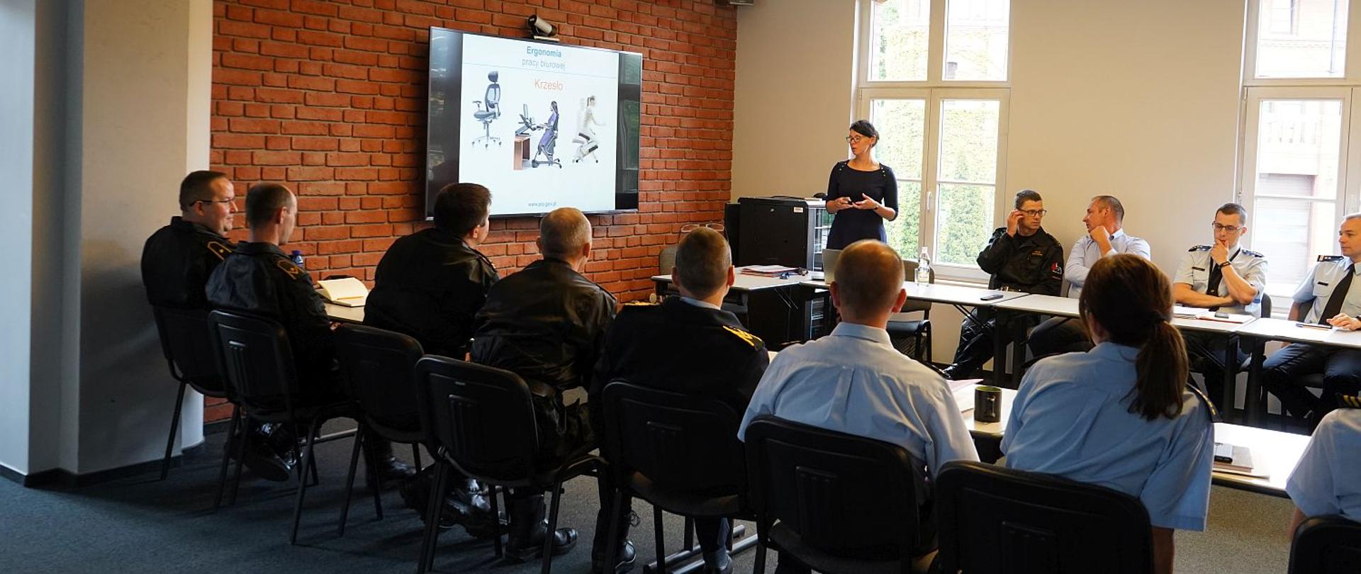 strażacy siedzący przy stołach, słuchajacy pani podczas szkolenia, na jeden jze ścian wisi telewizor