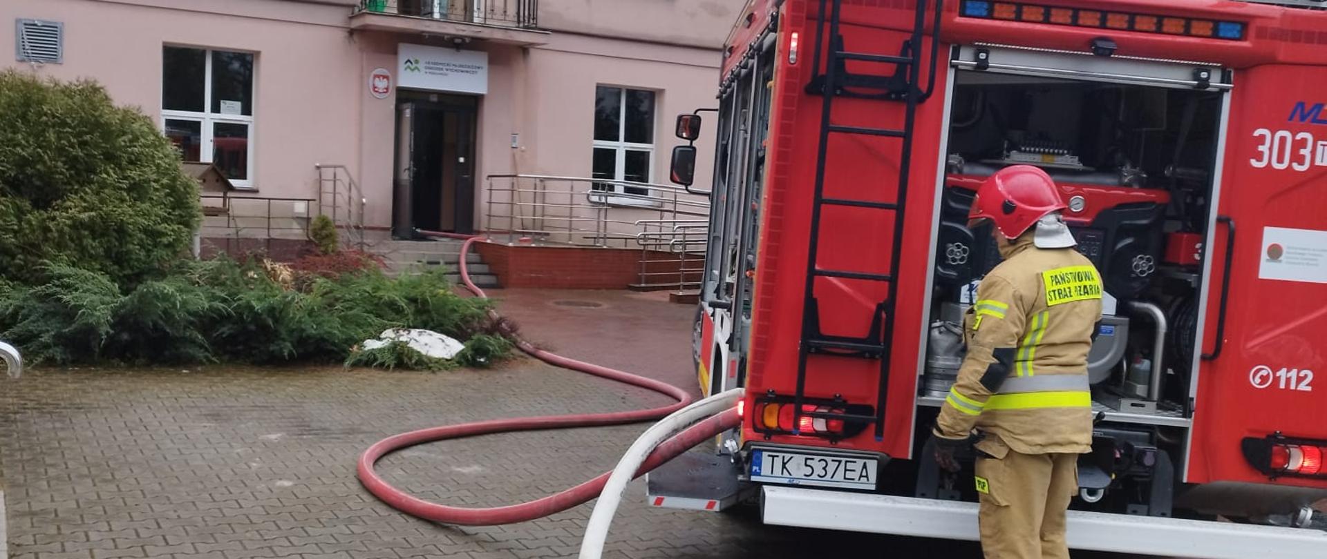 Zdjęcie przedstawia budynek ośrodka wychowawczego gdzie doszło do pożaru. Przed budynkiem stoi samochód pożarniczy, do którego podpięty jest wąż gaśniczy wchodzący do wnet raz obiektu. Przy samochodzie stoi strażak.