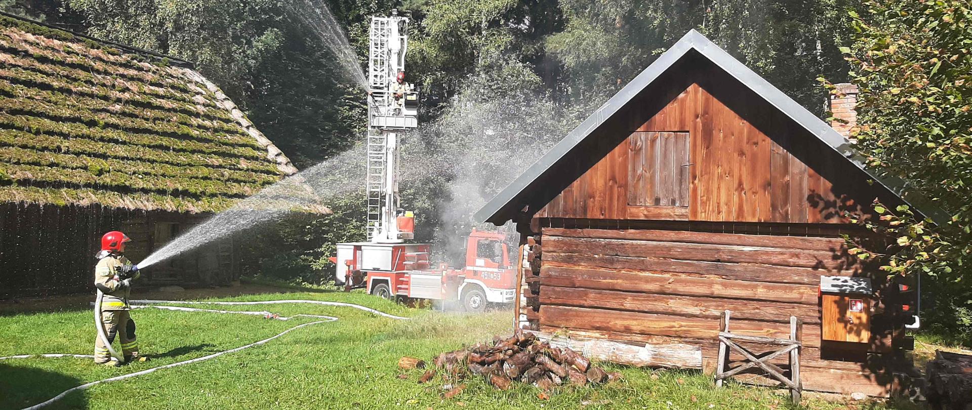 Na zdjęciu po lewej stronie część starego budynku krytego strzechą porośniętą mgłą, przed budynkiem stoi strażak w umundurowaniu bojowym, podaje jeden prąd wody. Po prawej stronie stary drewniany budynek. Pomiędzy budynkami węże strażackie oraz podnośnik hydrauliczny, z kosza którego podawany jest prąd wody. W tle drzewa.