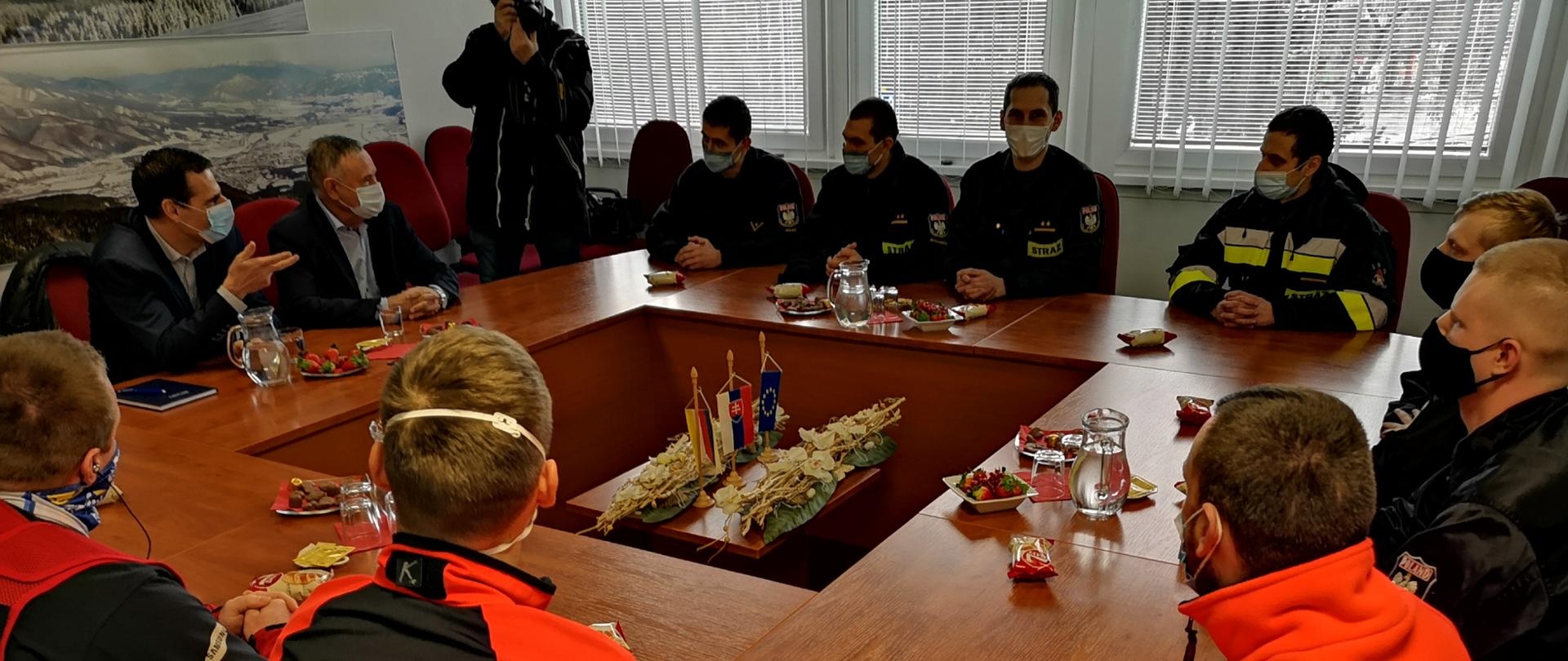 Wokół dużego kwadratowego stołu siedzą strażacy, ratownicy medyczni i cywile. Wszyscy w maseczkach. Po lewej stronie z tyłu stoi osoba robiąca zdjęcie aparatem fotograficznym. Trwa odprawa. Na środku stołu widoczne flagi Słowacji i Unii Europejskiej
