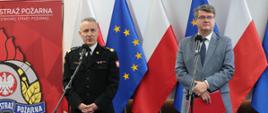 Mężczyzna w strażackim mundurze generalskim oraz drugi w okularach i szarym garniturze stojący przed mikrofonami. Za nim flagi Polski oraz Unii Europejskiej oraz logo Państwowej Straży Pożarnej