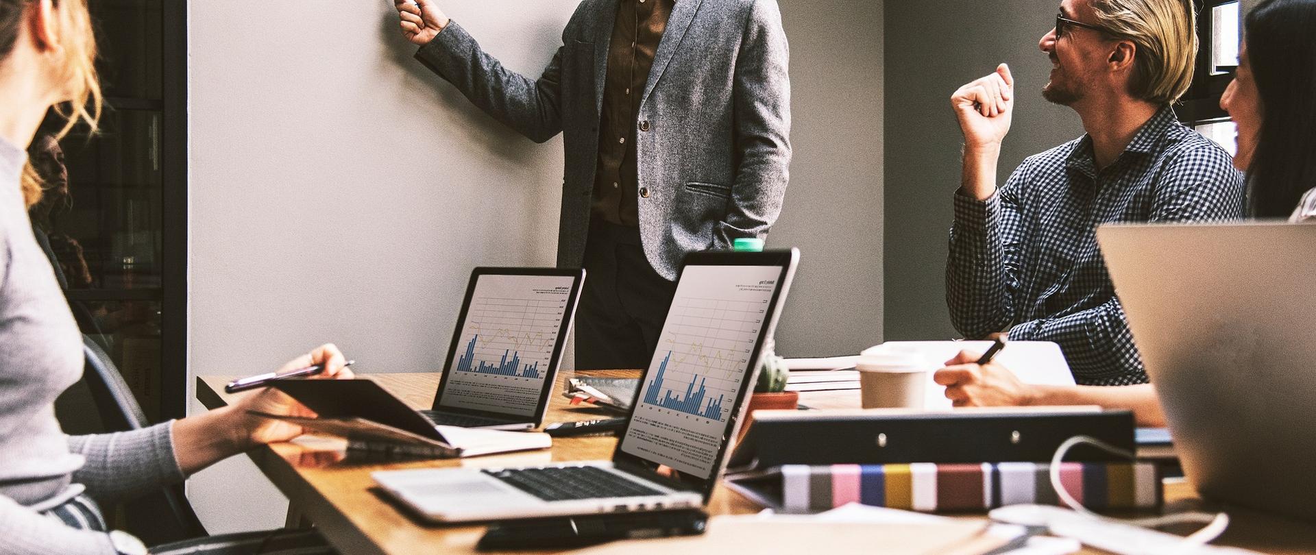 Spotkanie biznesowe, dwie osoby siedzą przy stołach konferencyjnych, jedna osoba wskazuje na tablicę