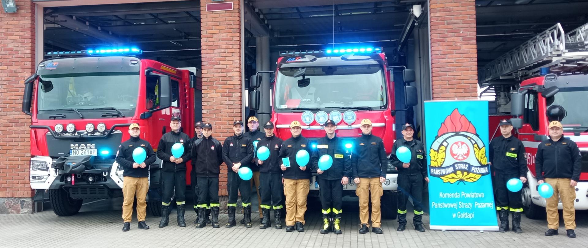 Zdjęcie przedstawia dwunastu funkcjonariuszy Komendy Powiatowej Państwowej Straży Pożarnej w Gołdapi wyposażonych w niebieskie akcenty (balony) na tle pojazdów pożarniczych z włączonymi niebieskimi sygnałami świetlnymi, solidarnych z osobami ze spektrum autyzmu