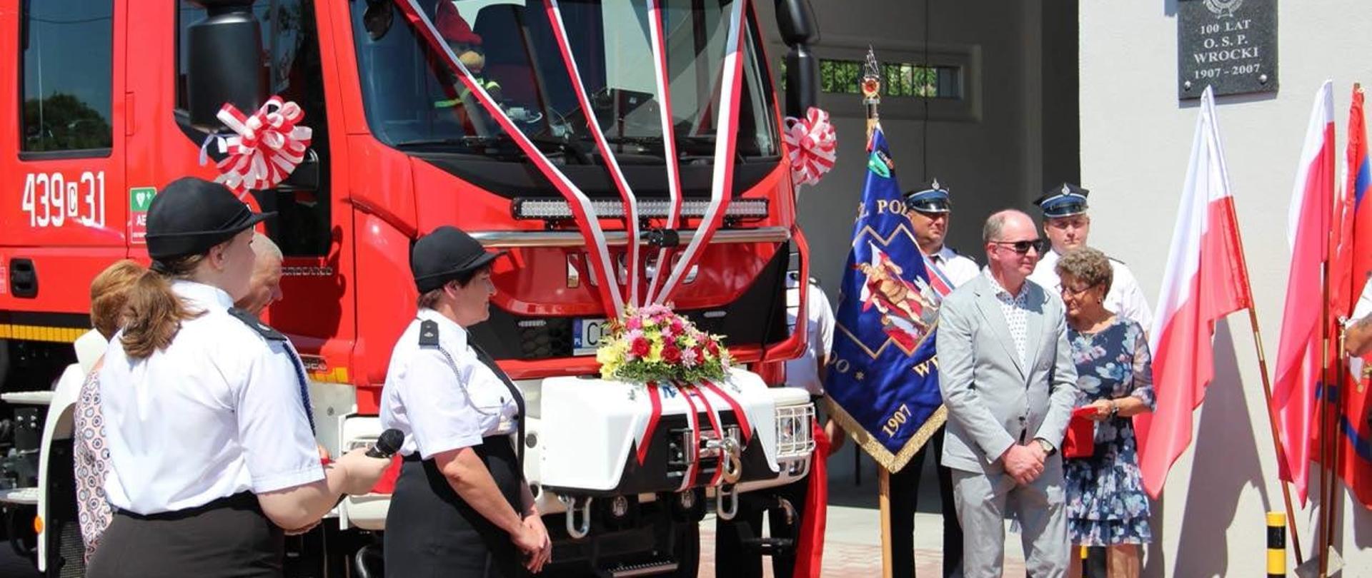Na zdjęciu druhny w umundurowaniu wyjściowym, stoją przy nowym aucie pożarniczym , udekorowanym odświętnie.W tle poczet sztandarowy.