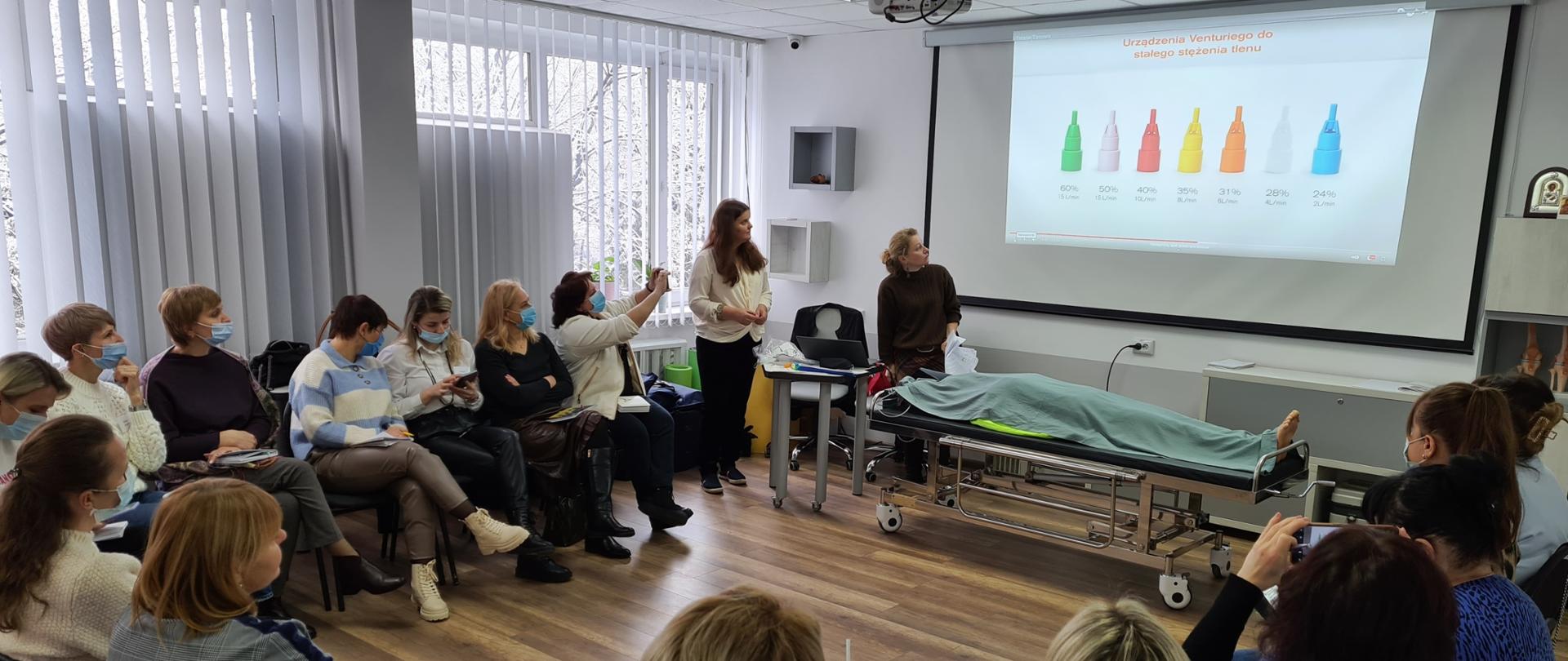 Kobiety siedzą w kręgu, obserwują prowadzącą, ekran i fantoma ratowniczego.