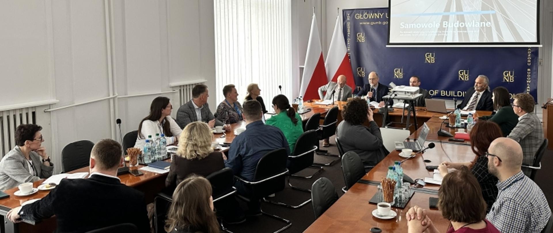 „Formalne spotkanie z prezentacją na temat samowoli budowlanych, logo GUNB, flaga Polski, uczestnicy przy stole z dokumentami i laptopami.”