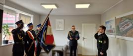 Zbiórka strażaków w jasnym pomieszczeniu. Po lewej stronie widoczny biało – czerwony - niebieski sztandar komendy. Po lewej funkcjonariusz w mundurze koszarowym składa ślubowanie. W środku komendant w mundurze wyjściowym odczytuje rotę ślubowania z granatowej teczki. W tle wyposażenie gabinetu.