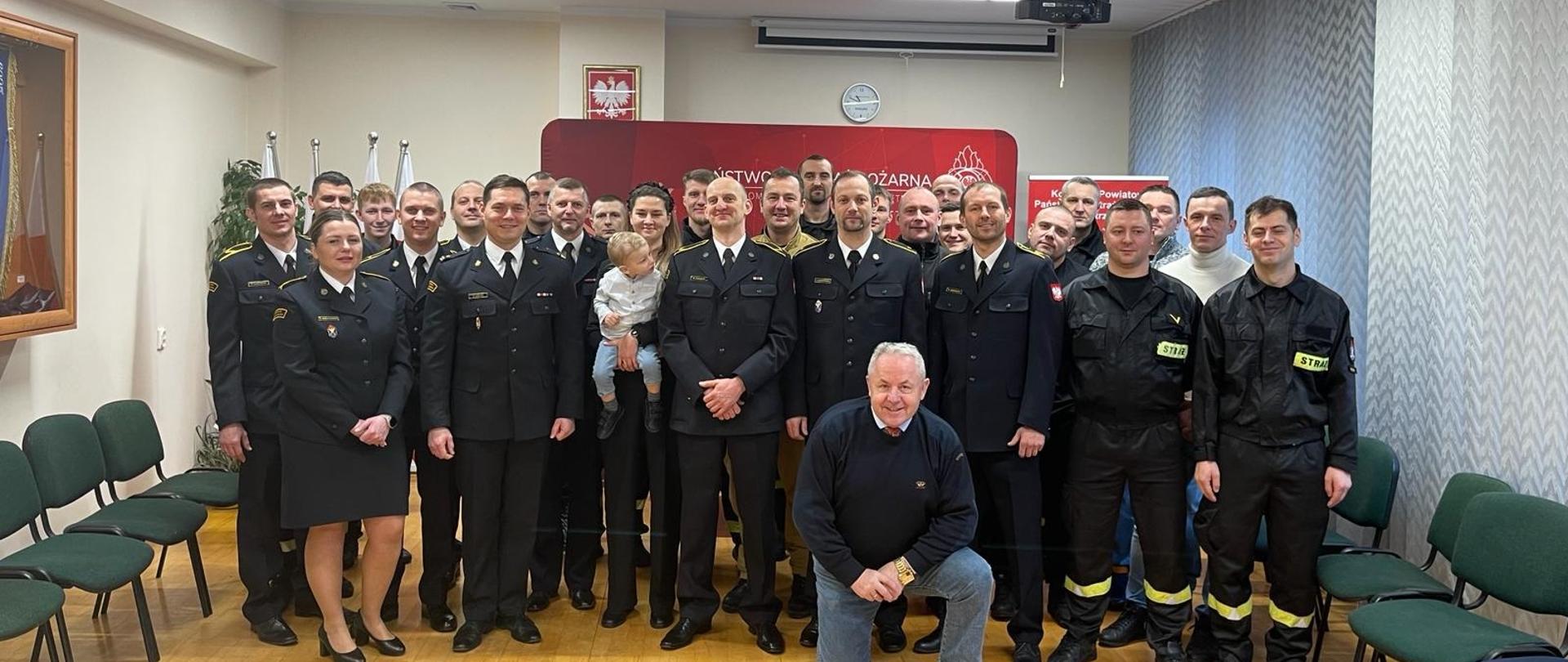 Zdjęcie grupowe na którym znajdują się odchodzący na zaopatrzenie emerytalne ogn. Wojciech Łuszczak z rodziną, Komendant Powiatowy i funkcjonariusze kętrzyńskiej Komendy oraz goście.