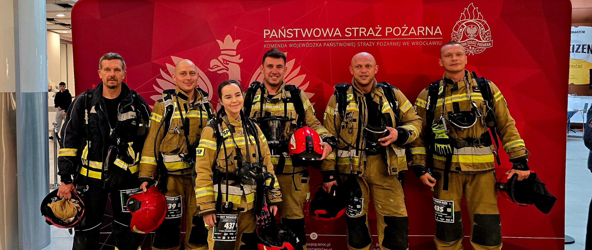 Strażacy w pełnym rynsztunku w budynkuGłówny plan / Osoby:Na zdjęciu stoi pięcioro strażaków – cztery osoby w drugim rzędzie oraz jedna kobieta wysunięta nieco do przodu. Wszystkie osoby są w pełnym umundurowaniu bojowym (piaskowe oraz czarne kurtki i spodnie strażackie z odblaskowymi żółtymi taśmami). Strażacy mają na sobie aparaty oddechowe z butlami na plecach, a na klatkach piersiowych przypięte papierowe numery startowe (m.in. z numerem 437, 39 oraz 135). Cześć osób trzyma w rękach czerwone hełmy strażackie. Wszyscy są uśmiechnięci i pozują przodem do aparatu. Tło i otoczenie:W tle znajduje się duża, czerwona ścianka reklamowa z godłem Polski (orzeł w koronie) oraz nagłówkiem: „PAŃSTWOWA STRAŻ POŻARNA - KOMENDA WOJEWÓDZKA PAŃSTWOWEJ STRAŻY POŻARNEJ WE WROCŁAWIU”. Podłoga wykonana jest z jasnych, połyskujących płytek ceramicznych, w których odbijają się postacie. Nad ścianką widać fragments koron drzew / roślin dekoracyjnych wewnątrz budynku oraz jasny hol. 
