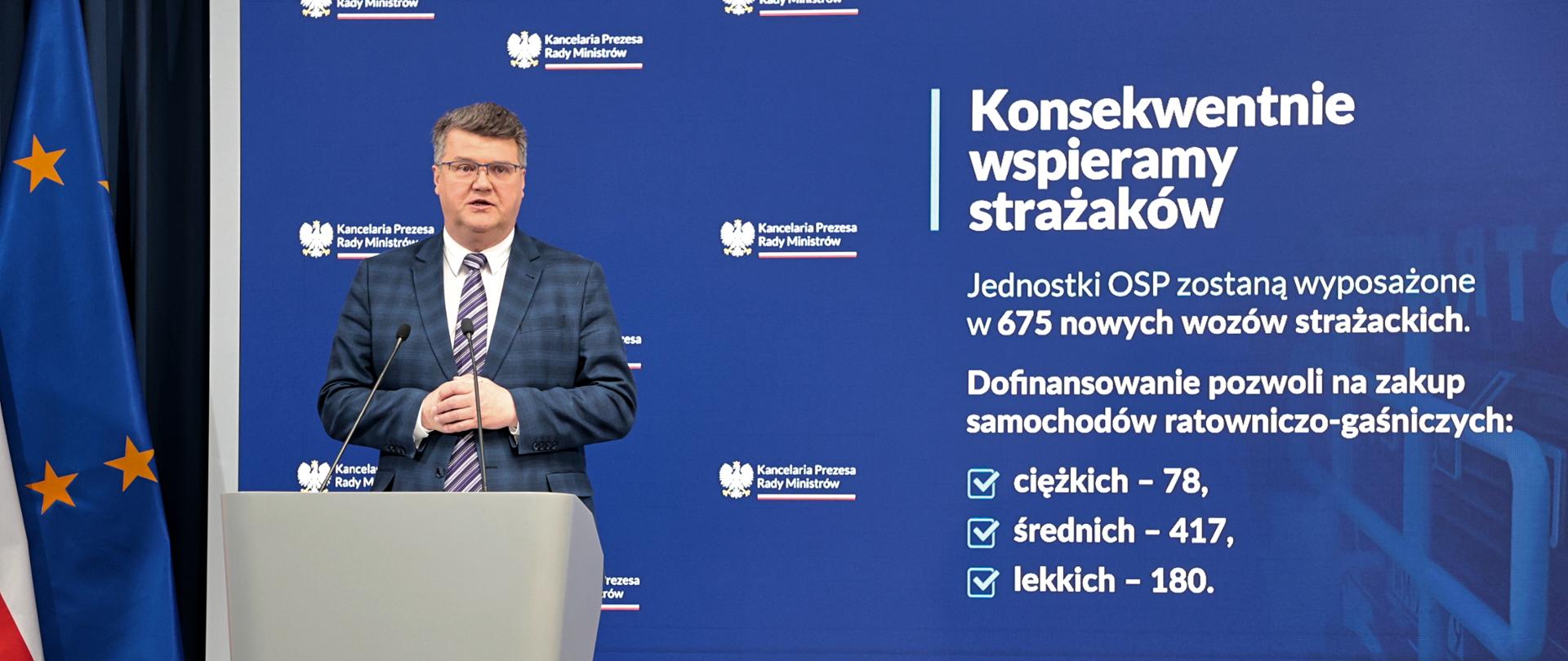 Maciej Wąsik przemawia przy mównicy w trakcie konferencji prasowej za nim banner mswia i napisami konsekwentnie wspieramy strażaków. Jednostki OSP zostaną wyposażone w 675 nowych wozów strażackich. dofinansowanie pozwoli na zakup samochodów ratowniczo-gaśniczych ciężkich 78 średnich 417 lekkich 180