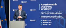Maciej Wąsik przemawia przy mównicy w trakcie konferencji prasowej za nim banner mswia i napisami konsekwentnie wspieramy strażaków. Jednostki OSP zostaną wyposażone w 675 nowych wozów strażackich. dofinansowanie pozwoli na zakup samochodów ratowniczo-gaśniczych ciężkich 78 średnich 417 lekkich 180