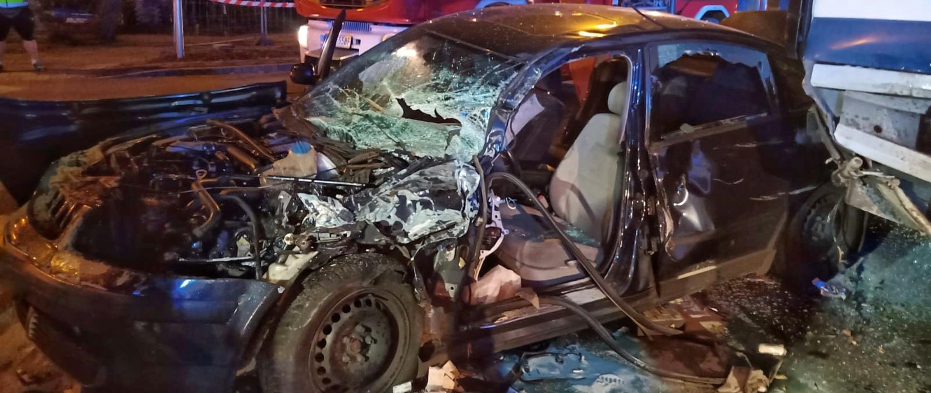 Na pierwszym planie widać bok auta osobowego po wypadku, bez maski, ze zbitą szybą przednią i bez drzwi bocznych. 