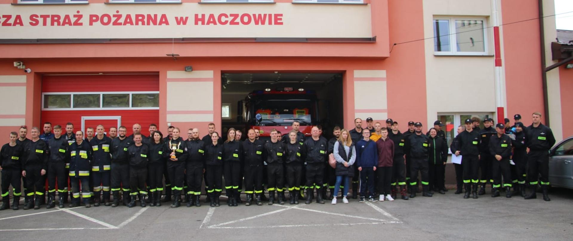 Kolorowa fotografia wykonana na zewnątrz w pogodny dzień. Przedstawia strażaków OSP w ubraniach koszarowych podczas podsumowania ćwiczeń „Haczów 2024” na tle remizy OSP w Haczowie.