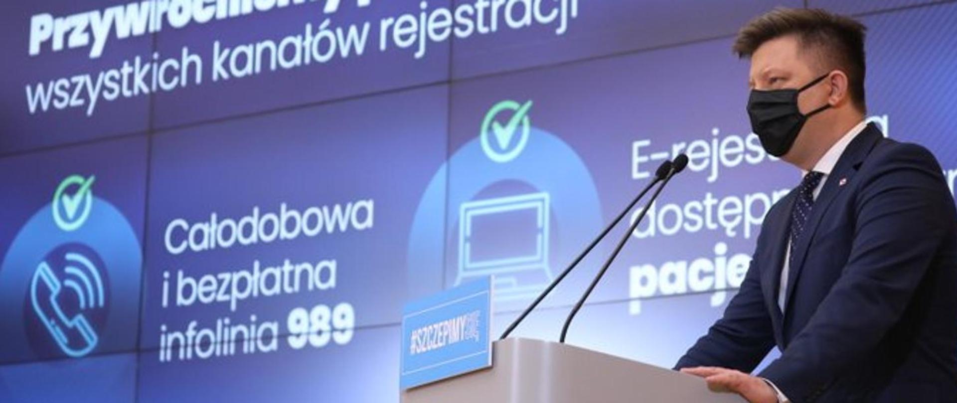 Na mównicy stoi mężczyzna w masce w tle widać całodobową infolinie 989 oraz rejestracje na szczepienia w e-rejestracji.