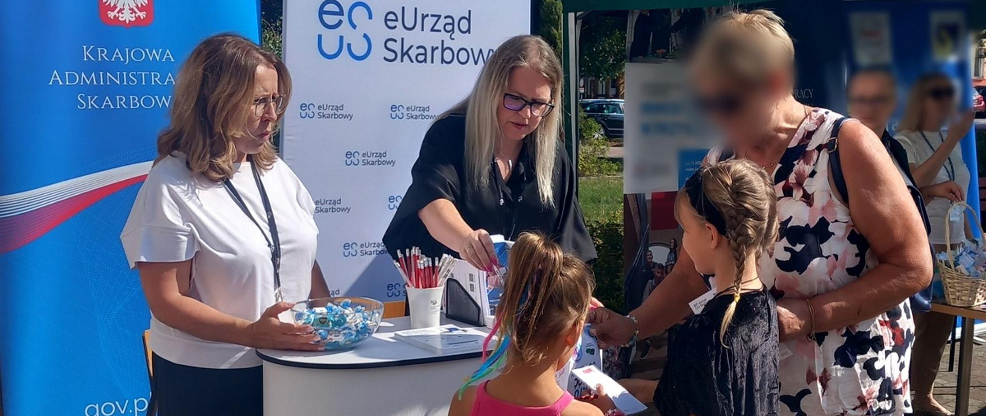 Stanowisko informacyjne KAS podczas pikniku w Tarnobrzegu. Ekspertki z Urzędu Skarbowego rozmawiają z odwiedzającymi stanowisko. W tle baner eUS i KAS.
