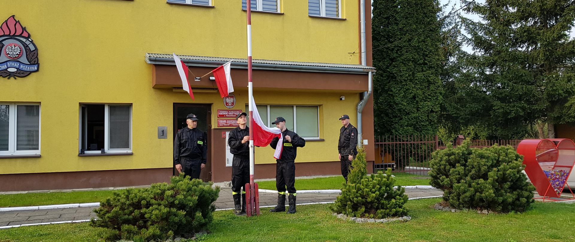Poczet flagowy składający się z trzech strażaków przygotowuje flagę państwową do podniesienie. W tle komendant powiatowy PSP w Proszowicach.