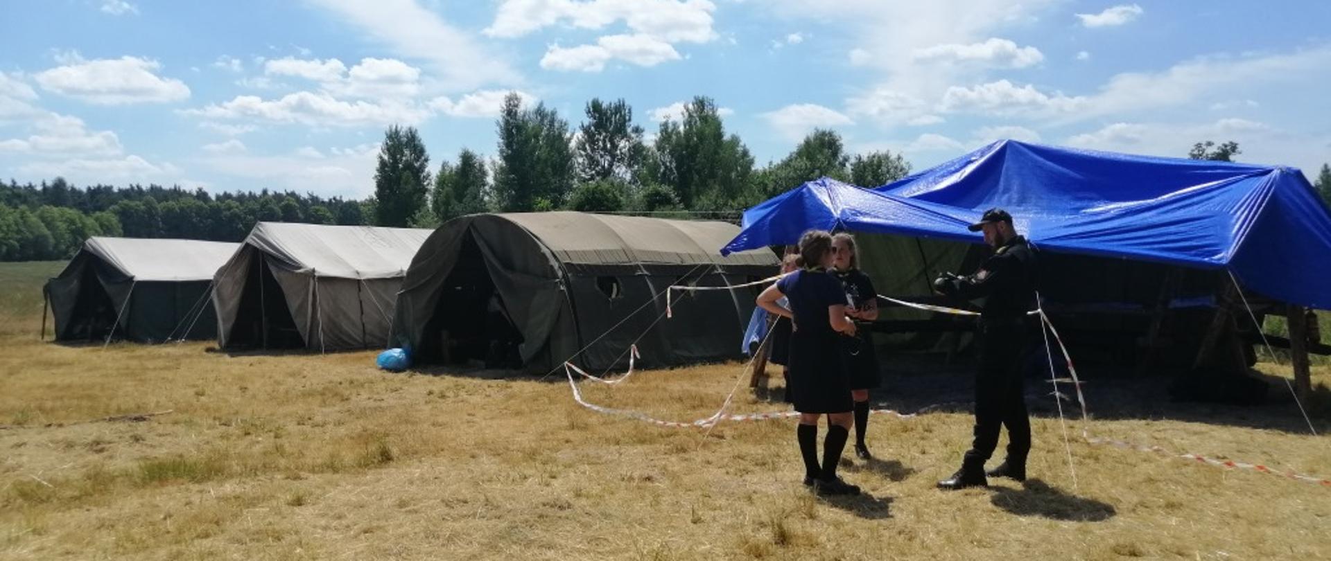 Zdjęcie przedstawia obóz harcerski znajdujący się na polanie. Na zdjęciu od lewej widać ustawione trzy namioty w kolorze ciemnym zielonym. Obok nich po prawej stronie znajduje się rozłożone zadaszenie chroniące przed słońcem wykonane z niebieskiej plandeki. Przed tym zadaszeniem stoją dwie harcerki oraz funkcjonariusz Państwowej Straży Pożarnej. Strażak dokonuje kontroli bezpiecznego użytkowania obozowiska. 