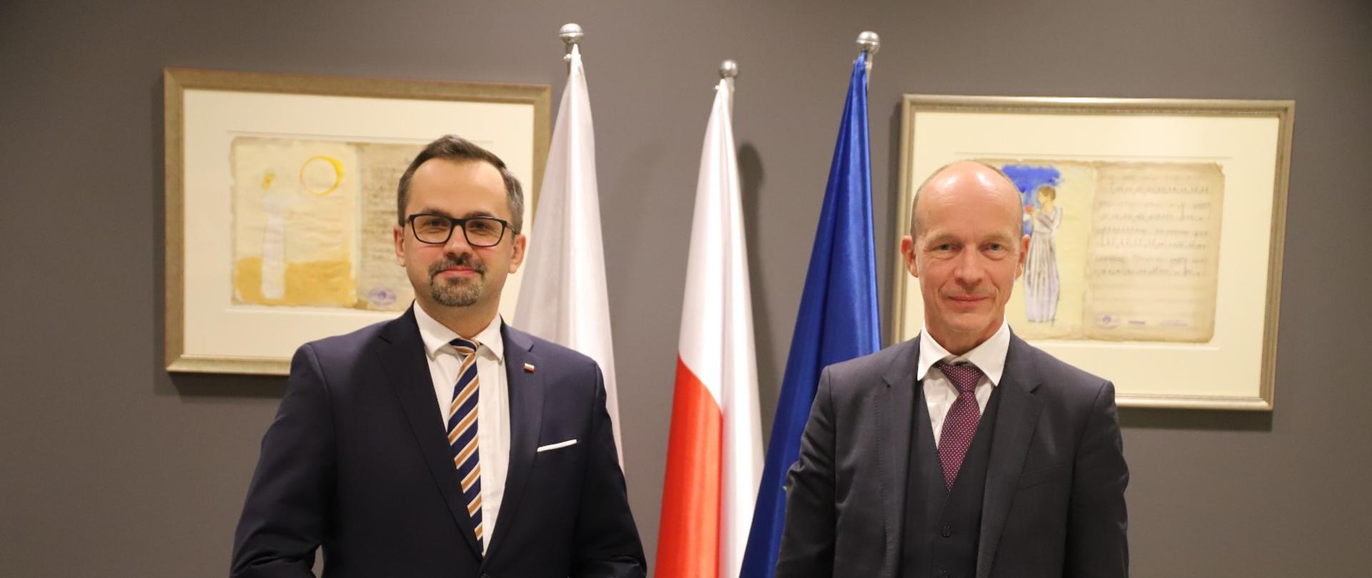 Dwie osoby stoją obok siebie. Pierwszy z lewej stoi wiceminister Marcin Horała. W tle stoją flagi Polski i Unii Europejskiej. 