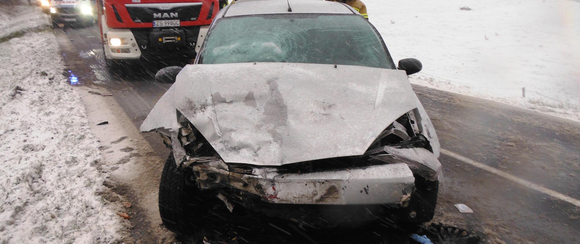 Na jezdni srebrny samochód osobowy z uszkodzonym przodem, samochód strażacki i karetka pogotowia. Padający śnieg.
