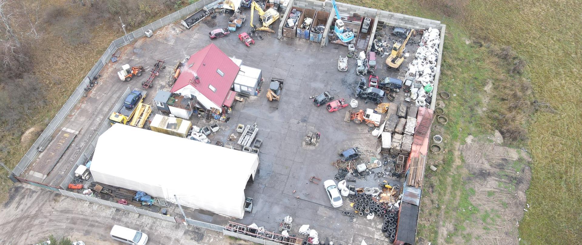 widok z góry: widoczny ogrodzony teren z budynkami i halą. Dookoła pojemniki i kontenery ze zdemontowanymi częściami, zdemontowane pojazdy, przenośniki dźwigowe,