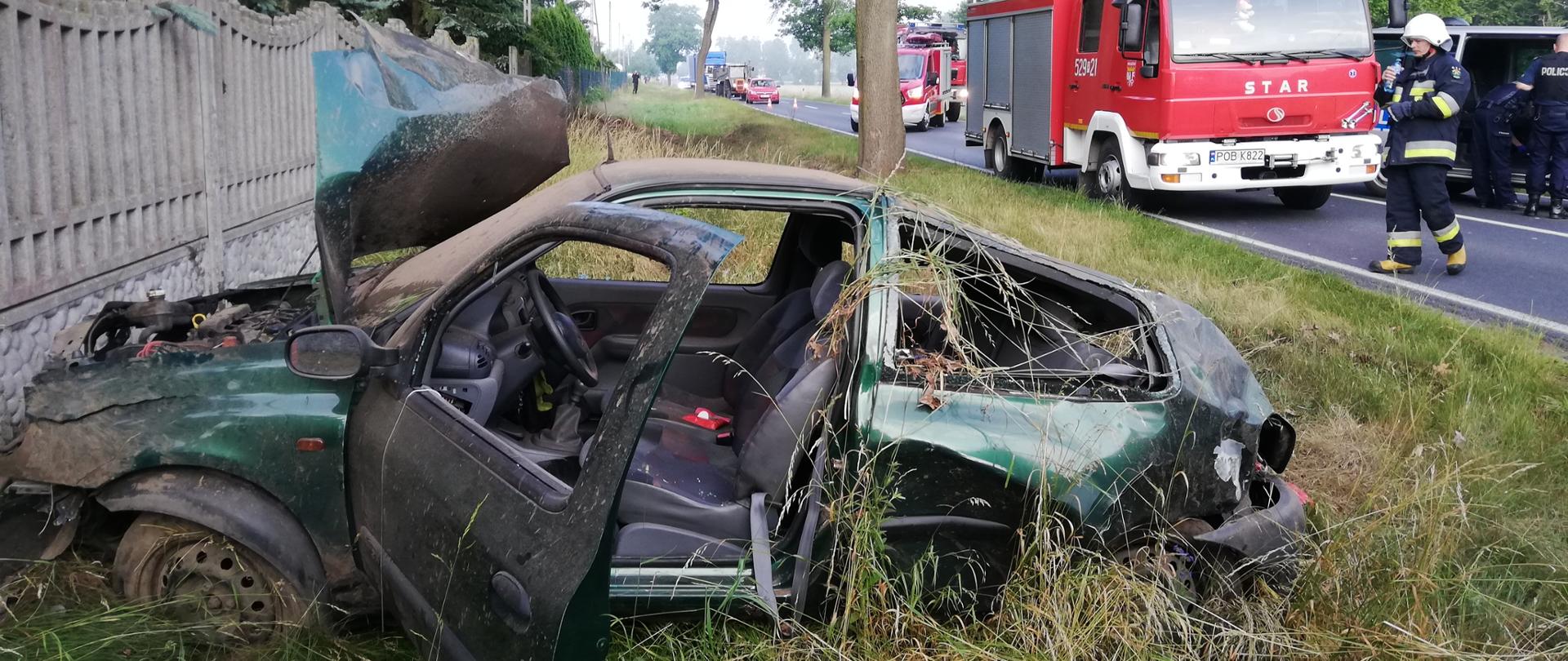 Na zdjęciu na pierwszym planie widoczny jest samochód osobowy marki Renault Clio po dachowaniu. Pojazd znajduje na poboczu drogi przy posesji. Na drugim planie widoczne pojazdy straży pożarnej oraz policji.