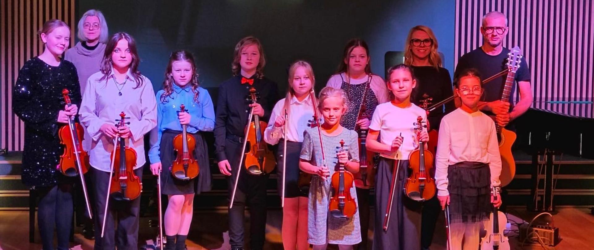 Dzieci pozują do zdjęcia ze skrzypcami. Stoją na scenie sali koncertowej. W tle widać ściane w kolorze zielonym oraz fragment fortepianu.