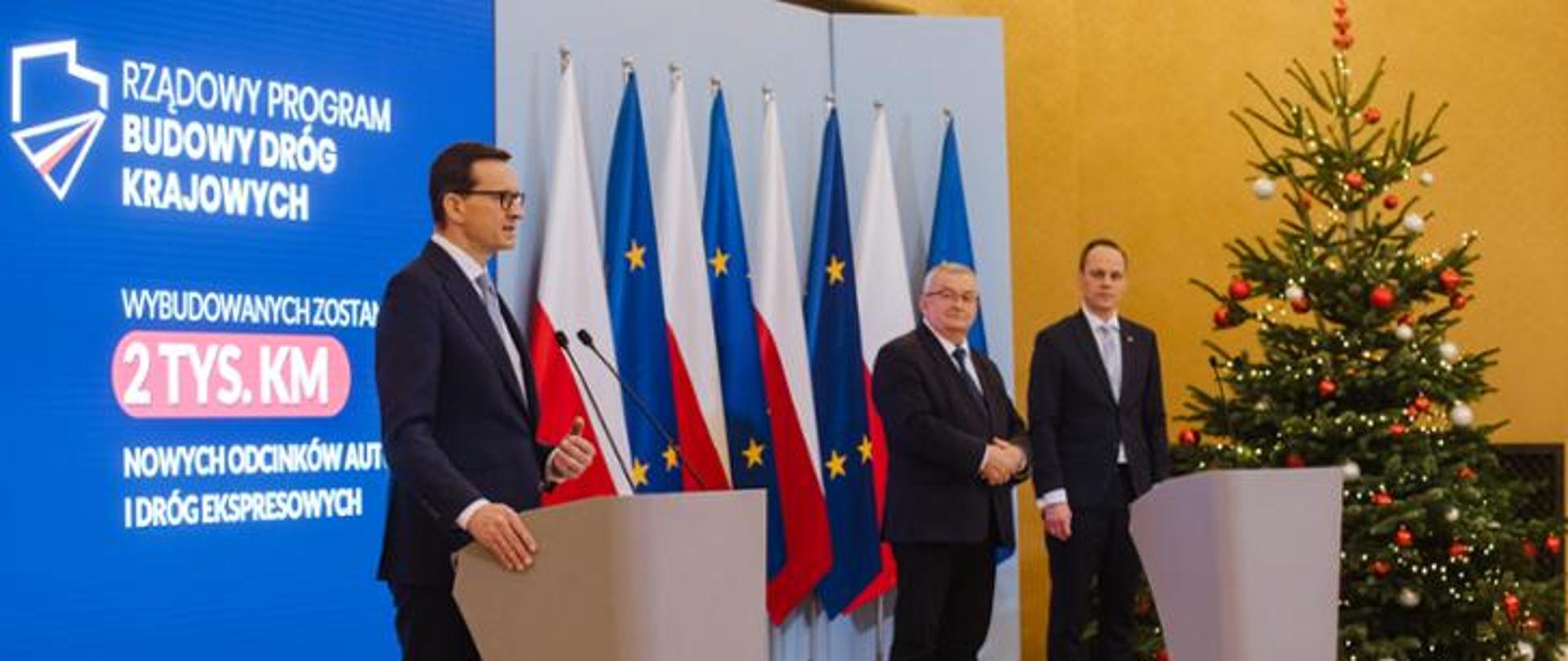 Jeden mężczyzna stojący przy mównicy i wypowiadający się. Obok stoi dwóch mężczyzn