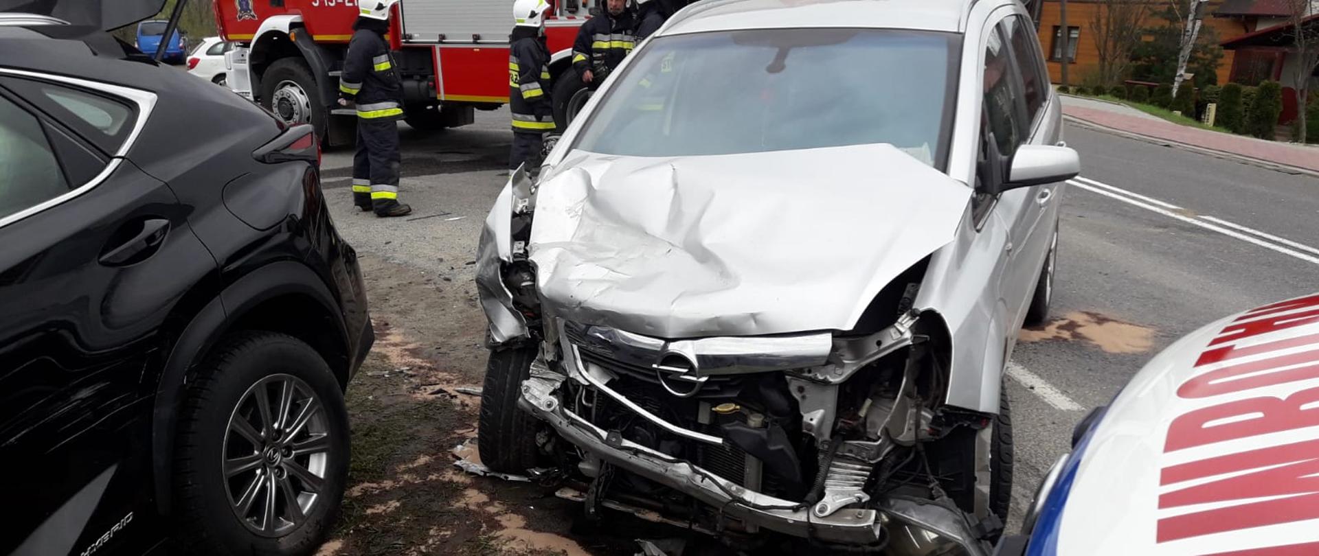 Widać tył samochodu osobowego ciemnego koloru oraz przód samochodu jasnego koloru uczestniczącego w zdarzeniu. Fragment karetki pogotowia ratunkowego oraz samochodu pożarniczego.